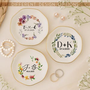 May include: Three white ceramic ring dishes with gold rims, each featuring a floral wreath design and personalized text. The text includes initials and dates. The image also shows a pearl bracelet, rings, and the text "DIFFERENT DESIGN OPTIONS".
