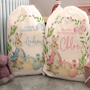 Pu&ograve; includere: Due sacchetti regalo di Pasqua in tela bianca con nomi personalizzati. Il sacchetto a sinistra ha un coniglio blu con uova blu e il testo "Easter Delivery for Andrew". Il sacchetto a destra ha un coniglio rosa con uova rosa e il testo "Easter Delivery for Chloe".