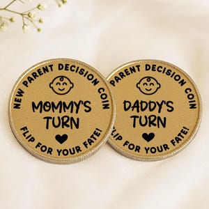 May include: Two gold-colored decision coins with black text. One coin reads "Mommy's Turn" and the other reads "Daddy's Turn". Both coins have a baby face icon and the phrase "Flip for your fate!" around the edge.