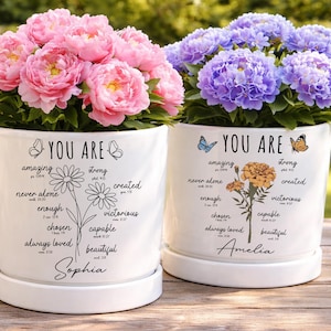 May include: Two white ceramic flower pots with floral arrangements. One pot has pink flowers, the other has purple flowers. Each pot has the words "YOU ARE" and inspirational words. The first pot is labeled "Sophia", the second "Amelia".