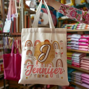 K&ouml;nnte beinhalten: Eine wei&szlig;e Canvas-Tragetasche mit einem bunten Regenbogen-Design und dem Namen "Jennifer" in Pink bedruckt. Die Tasche hat au&szlig;erdem einen gro&szlig;en Buchstaben "J" in einem goldenen Kreis.