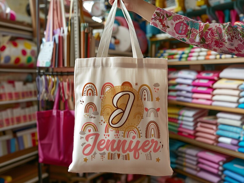 K&ouml;nnte beinhalten: Eine wei&szlig;e Canvas-Tragetasche mit einem bunten Regenbogen-Design und dem Namen "Jennifer" in Pink bedruckt. Die Tasche hat au&szlig;erdem einen gro&szlig;en Buchstaben "J" in einem goldenen Kreis.