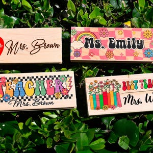 K&ouml;nnte beinhalten: Vier Namensschilder f&uuml;r Lehrer aus Holz mit verschiedenen Designs. Eines hat einen roten Apfel und die Worte "Teacher life" und "Mrs. Brown". Ein anderes zeigt einen Regenbogen und den Namen "Ms. Emily". Das dritte sagt "Teacher Mrs. Brown". Das letzte sagt "Best Teacher Mrs. Watson".
