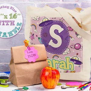 K&ouml;nnte beinhalten: Eine wei&szlig;e Canvas-Tragetasche mit einem lila und pinkfarbenen Design mit einem gro&szlig;en Buchstaben "S" und dem Wort "Sarah". Die Tasche hat eine Tragetaschengr&ouml;&szlig;e von 38 cm x 41 cm mit 25 cm langen Griffen.