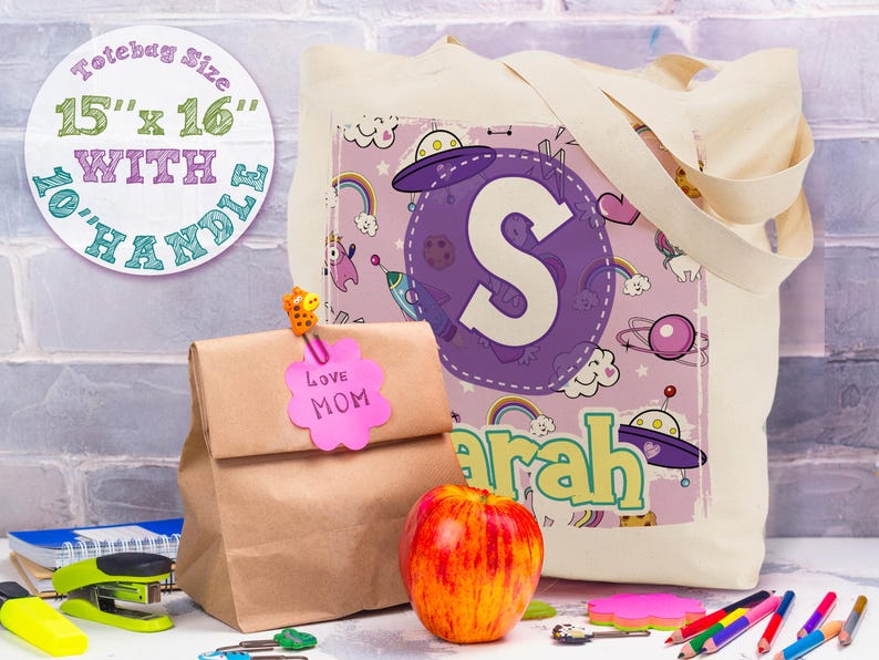 K&ouml;nnte beinhalten: Eine wei&szlig;e Canvas-Tragetasche mit einem lila und pinkfarbenen Design mit einem gro&szlig;en Buchstaben "S" und dem Wort "Sarah". Die Tasche hat eine Tragetaschengr&ouml;&szlig;e von 38 cm x 41 cm mit 25 cm langen Griffen.