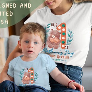 Puede incluir: Una camiseta blanca y un body azul claro con una ilustraci&oacute;n de oso y el texto "Our 1st Mother's Day", adem&aacute;s de los nombres "Huy & Kevin 2026". Tambi&eacute;n es visible el texto "Designed and Printed in USA".