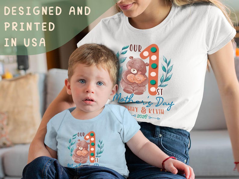 Puede incluir: Una camiseta blanca y un body azul claro con una ilustraci&oacute;n de oso y el texto "Our 1st Mother's Day", adem&aacute;s de los nombres "Huy & Kevin 2026". Tambi&eacute;n es visible el texto "Designed and Printed in USA".