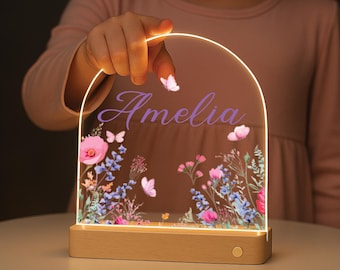 May include: A personalized night light with the name "Amelia" in purple script. The clear acrylic panel features a floral design with pink poppies, butterflies, and other flowers. The base is made of light-colored wood.