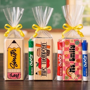 May include: Three gift bags with personalized wooden blocks and markers. One block features a pencil design with "Smith" and "Mrs." text. Another has an apple and ruler with "Ms. Thompson". The third has a rainbow and flowers with "Ms. Emily".