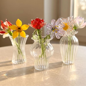May include: Three clear glass vases each hold a different colorful glass flower. The flowers include red, yellow, white, and purple blooms. The vases are on a light-colored surface.
