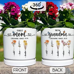 May include: White ceramic planter with a floral design and the text "First Mom Growing Since 1974" on the front and "Now Grandma Growing Since 2025" on the back. The planter has a saucer and is filled with green plants and pink flowers.