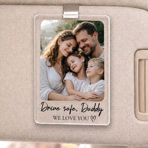 May include: A clear acrylic car visor photo holder with a metal clip. The holder displays a family photo and the text "Drive safe, Daddy WE LOVE YOU". The visor is attached to a beige car interior.