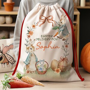 Pu&ograve; includere: Una borsa a cordoncino bianca con bordino rosso che presenta un'illustrazione ad acquerello di due conigli che tengono carote e uova di Pasqua. La borsa ha il testo "Easter Delivery for Sophia" stampato su di essa.