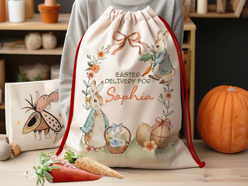 Pu&ograve; includere: Una borsa a cordoncino bianca con bordino rosso che presenta un'illustrazione ad acquerello di due conigli che tengono carote e uova di Pasqua. La borsa ha il testo "Easter Delivery for Sophia" stampato su di essa.