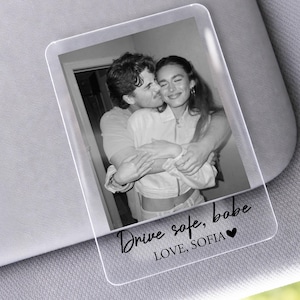 May include: Clear acrylic car visor photo with a black and white image of a couple embracing. The text "Drive safe, babe" and "LOVE, SOFIA" is printed in black script with a heart symbol.