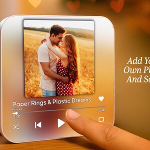 May include: A silver-colored digital music player with a touch screen displays a photo of a couple embracing. The screen shows the text "Paper Rings & Plastic Dreams" and playback controls. The text "Add Your Own Photo And Song" is also visible.