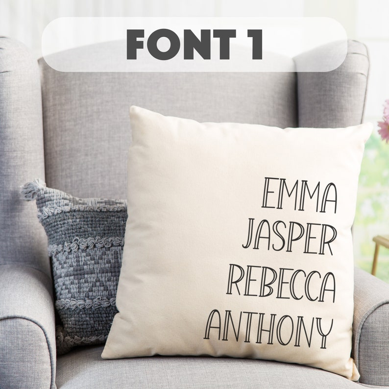 May include: A white pillow with black text that reads "EMMA JASPER REBECCA ANTHONY" in a decorative font. The pillow is sitting on a gray armchair.
