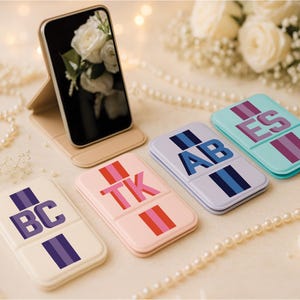 May include: A smartphone displays a floral image, propped on a beige stand. Four rectangular cases with initials and striped accents are arranged below. The cases are in cream, pink, lavender, and teal, with pearl strands and flowers in the background.