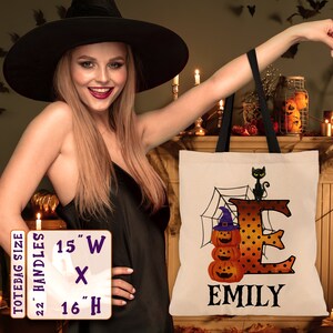 May include: A white tote bag with black handles. The bag features a black cat, a spiderweb, and a large orange letter "E" with black polka dots. The letter "E" is decorated with three orange pumpkins. The name "EMILY" is printed in black below the letter. The tote bag measures 15 inches wide by 16 inches high.