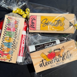 K&ouml;nnte beinhalten: Geschenksets f&uuml;r Lehrer, jeweils in Zellophan mit gelbem Band verpackt. Ein Set enth&auml;lt Marker und einen Holzblock mit dem Wort TEACHER. Ein weiteres zeigt ein Bleistift-Design mit dem Namen Smith. Das dritte Set hat die Worte #TeacherLife.