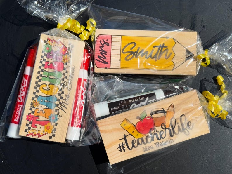 K&ouml;nnte beinhalten: Geschenksets f&uuml;r Lehrer, jeweils in Zellophan mit gelbem Band verpackt. Ein Set enth&auml;lt Marker und einen Holzblock mit dem Wort TEACHER. Ein weiteres zeigt ein Bleistift-Design mit dem Namen Smith. Das dritte Set hat die Worte #TeacherLife.