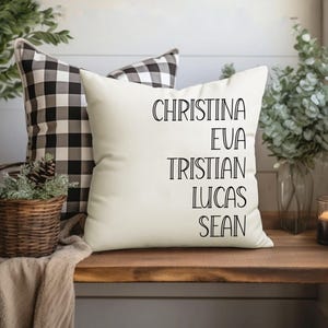 May include: White pillow with black text that reads "Christina, Eva, Tristian, Lucas, Sean". The pillow is on a wooden surface with a black and white checkered pillow behind it.