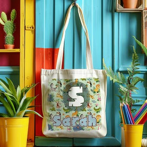 K&ouml;nnte beinhalten: Eine wei&szlig;e Canvas-Tragetasche mit einem Dschungeltier-Druck und dem Namen "Sarah" in einer bunten Schrift. Die Tasche hat einen gro&szlig;en Buchstaben "S" in einem Kreis auf der Vorderseite.