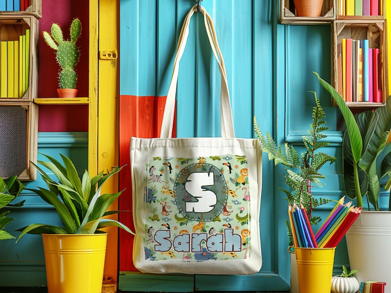 K&ouml;nnte beinhalten: Eine wei&szlig;e Canvas-Tragetasche mit einem Dschungeltier-Druck und dem Namen "Sarah" in einer bunten Schrift. Die Tasche hat einen gro&szlig;en Buchstaben "S" in einem Kreis auf der Vorderseite.