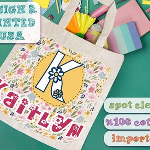K&ouml;nnte beinhalten: Eine wei&szlig;e Canvas-Tragetasche mit einem Blumenmuster und dem Namen "Kaitlyn" in Pink bedruckt. Der Buchstabe "K" ist in einem gelben Kreis mit blauem Rand hervorgehoben. Die Tasche ist aus 100% Baumwolle und importiert. Nur Fleckenreinigung.