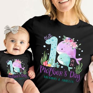 Puede incluir: Camisetas negras con un dise&ntilde;o a juego que presenta una ilustraci&oacute;n de ballena, flores y el texto "Our 1st Mother's Day". Una camiseta es para un adulto, la otra para un beb&eacute;. El dise&ntilde;o incluye el a&ntilde;o 2026 y los nombres "Mommy & Amelia."