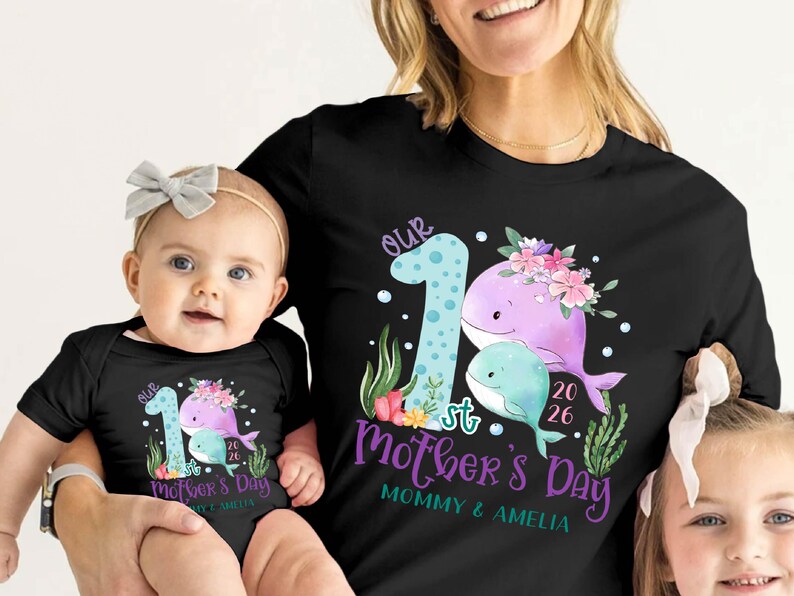 Puede incluir: Camisetas negras con un dise&ntilde;o a juego que presenta una ilustraci&oacute;n de ballena, flores y el texto "Our 1st Mother's Day". Una camiseta es para un adulto, la otra para un beb&eacute;. El dise&ntilde;o incluye el a&ntilde;o 2026 y los nombres "Mommy & Amelia."