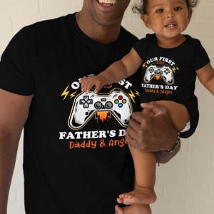 May include: A black t-shirt with a graphic of a video game controller and the text "Our First 2023 Father's Day Daddy & Angel". A child is wearing a matching black t-shirt with the text "Our First 2023 Father's Day Daddy & Angel".