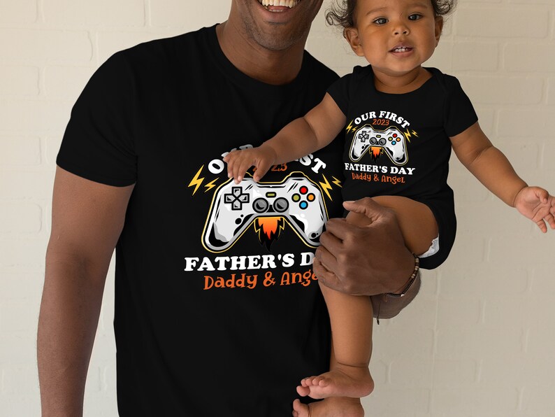 May include: A black t-shirt with a graphic of a video game controller and the text "Our First 2023 Father's Day Daddy & Angel". A child is wearing a matching black t-shirt with the text "Our First 2023 Father's Day Daddy & Angel".