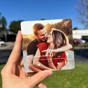 May include: A square, transparent acrylic photo print featuring a couple embracing, with the text "Through Every Storm" and a music player interface. The background shows a sunny outdoor scene.