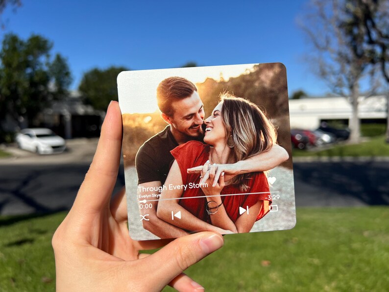 May include: A square, transparent acrylic photo print featuring a couple embracing, with the text "Through Every Storm" and a music player interface. The background shows a sunny outdoor scene.