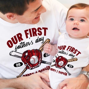 May include: A white t-shirt with the text "Our First Father's Day" in red lettering, featuring a baseball glove and bats. The shirt also includes the year "20" and the text "Daddy & Sophia". A baby is wearing a matching white onesie with the text "Our First Father's Day" in red lettering, featuring a baseball glove and bats. The onesie also includes the year "23" and the text "Daddy & Sophia".