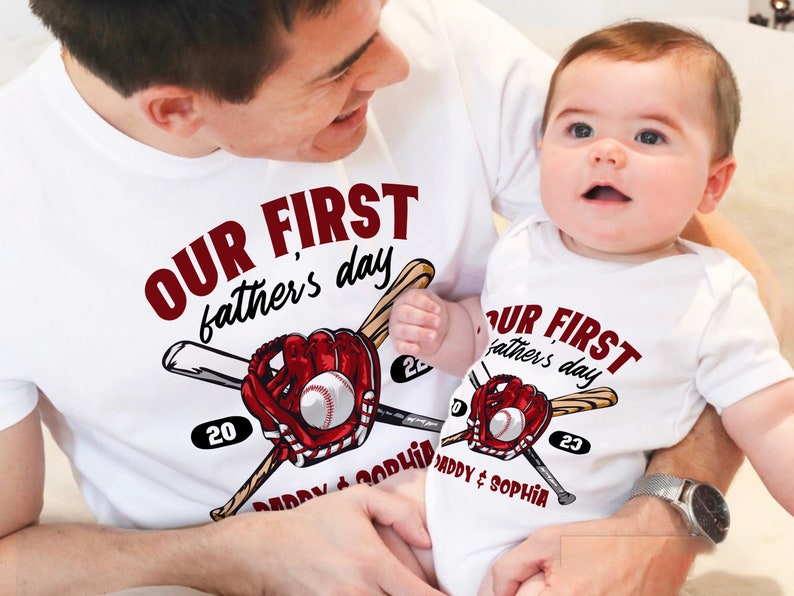 May include: A white t-shirt with the text "Our First Father's Day" in red lettering, featuring a baseball glove and bats. The shirt also includes the year "20" and the text "Daddy & Sophia". A baby is wearing a matching white onesie with the text "Our First Father's Day" in red lettering, featuring a baseball glove and bats. The onesie also includes the year "23" and the text "Daddy & Sophia".