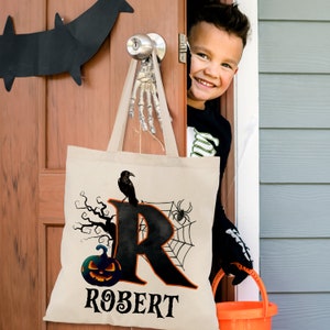 May include: A white canvas tote bag with a black and orange Halloween design. The bag features a black crow, a spider web, a pumpkin, and the letter "R" in black. The name "ROBERT" is printed in black at the bottom of the bag.