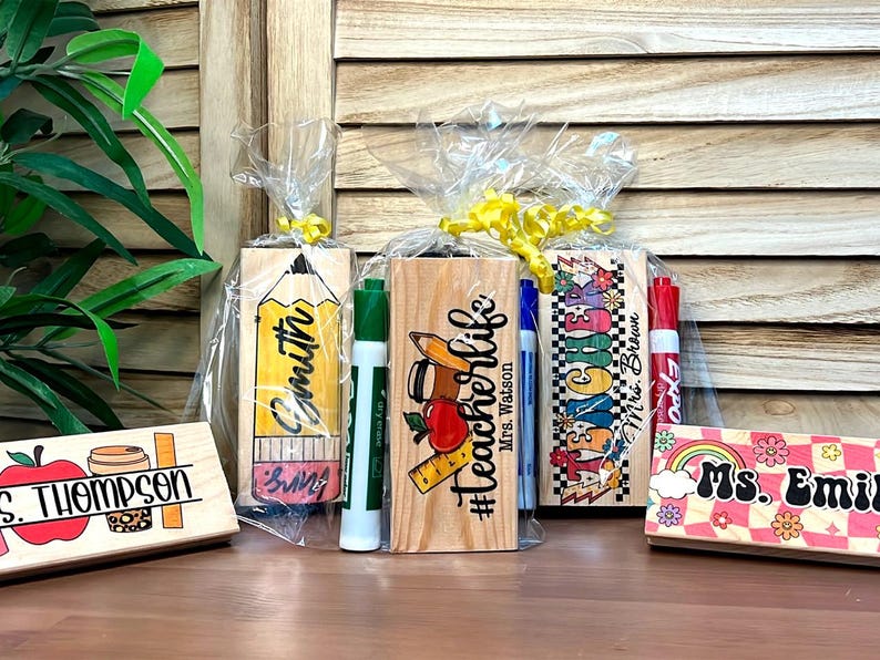 K&ouml;nnte beinhalten: Holz-Lehrerschreibtisch-Namensschilder mit farbenfrohen Designs und Lehrernamen. Eines zeigt einen Apfel und eine Kaffeetasse, ein anderes einen Bleistift und ein weiteres ein Lineal und einen Apfel. Jedes ist mit einer Klarsichtfolie und gelbem Band verpackt.