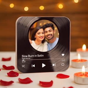 May include: A square, black and gold photo frame with a circular photo of a smiling couple. The frame has a vinyl record design and displays song information: "Slow Burn in Satin" by Tahila Monroe. Red rose petals and lit candles surround the frame.