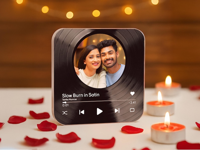 May include: A square, black and gold photo frame with a circular photo of a smiling couple. The frame has a vinyl record design and displays song information: "Slow Burn in Satin" by Tahila Monroe. Red rose petals and lit candles surround the frame.