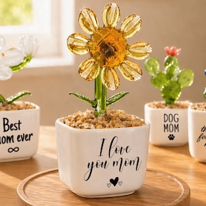 May include: A collection of decorative glass flower sculptures in white ceramic pots. The flowers include a yellow sunflower, a white flower, a green cactus, and a blue flower. Each pot has a different message, such as "I love you Mom."
