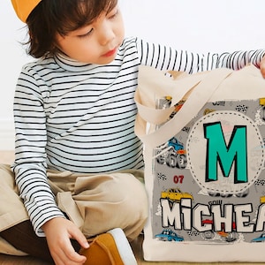 K&ouml;nnte beinhalten: Eine beige Canvas-Tragetasche mit Monstertruck-Design. Die Tasche hat einen gro&szlig;en t&uuml;rkisfarbenen Buchstaben "M" in einem Kreis und den Namen "MICHEAL" in wei&szlig;en Buchstaben. Die Tasche hat schwarze Henkel.