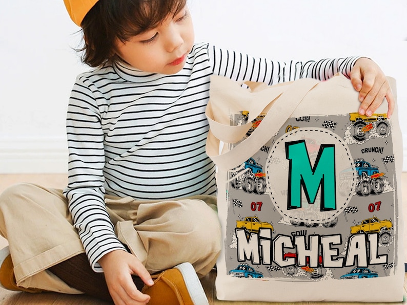K&ouml;nnte beinhalten: Eine beige Canvas-Tragetasche mit Monstertruck-Design. Die Tasche hat einen gro&szlig;en t&uuml;rkisfarbenen Buchstaben "M" in einem Kreis und den Namen "MICHEAL" in wei&szlig;en Buchstaben. Die Tasche hat schwarze Henkel.