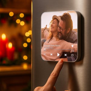 May include: A square, silver-framed digital photo frame displays a couple embracing, with the text "Through Every Storm." The frame shows a music player interface. A finger touches the frame. Red candles and bokeh lights are in the background.