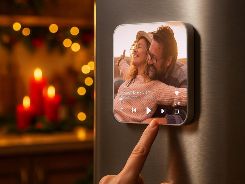 May include: A square, silver-framed digital photo frame displays a couple embracing, with the text "Through Every Storm." The frame shows a music player interface. A finger touches the frame. Red candles and bokeh lights are in the background.