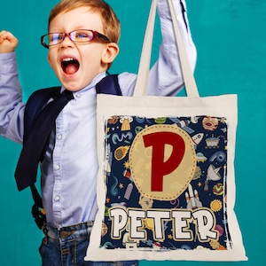 K&ouml;nnte beinhalten: Eine wei&szlig;e Canvas-Tragetasche mit einem blauen und gelben Weltraum-Design. Die Tasche hat einen gro&szlig;en roten Buchstaben "P" und den Namen "PETER" in wei&szlig; gedruckt.