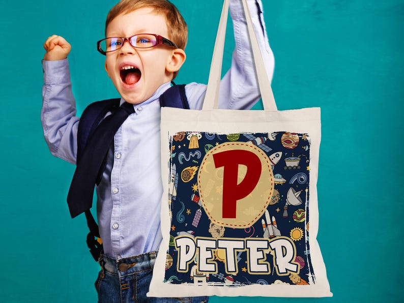 K&ouml;nnte beinhalten: Eine wei&szlig;e Canvas-Tragetasche mit einem blauen und gelben Weltraum-Design. Die Tasche hat einen gro&szlig;en roten Buchstaben "P" und den Namen "PETER" in wei&szlig; gedruckt.