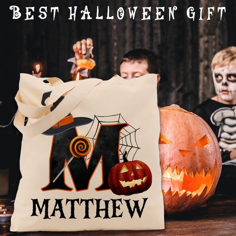 May include: A beige canvas tote bag featuring a black letter M, a witch's hat, a spiderweb, and a jack-o'-lantern. The bag is personalised with the name "MATTHEW".