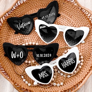 May include: A set of five heart-shaped sunglasses with black frames and lenses. The sunglasses are decorated with white text, including "Bride", "Victoria", "W+O", "10.10.2024", "Harris", and "Mrs."